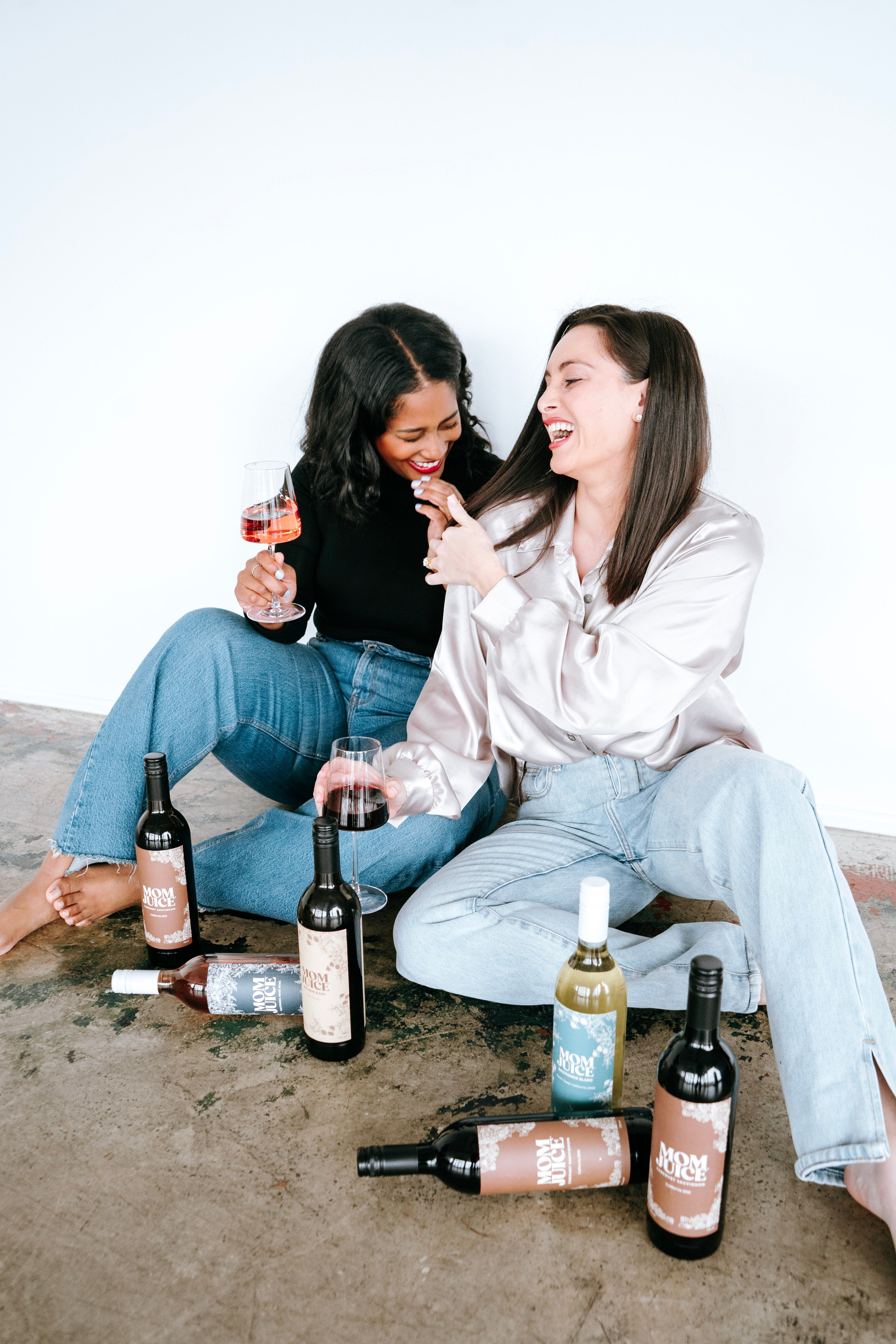 kristin and macie lifestyle image  on ground with all types of mom juice and their wine in hand