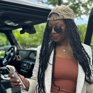 Black woman with locs and phone wearing Mom Juice hat in sand in front of G wagon