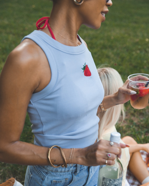 Woman in light blue sleeveless top with strawberry design, holding a drink outdoors.