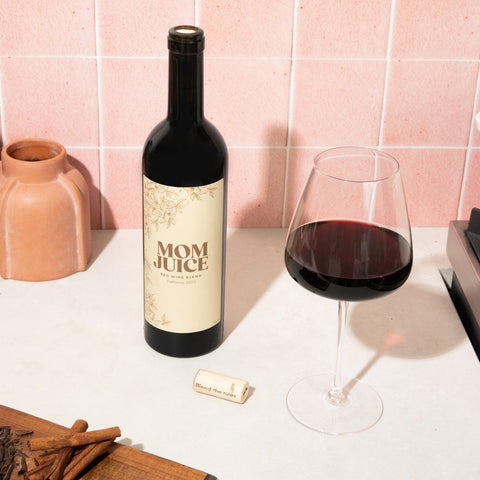 Downward angle mom juice red blend shot with full glass of wine, wood plank, cinnamon, choclate, pink tile and vase