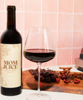 Mom Juice Red Blend on Pink tile background with wood plank, spices, wine glass